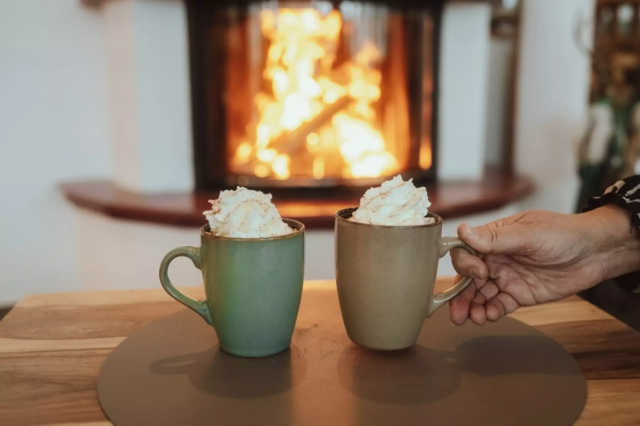 Zwei Tassen heiße Schokolade mit Sahne stehen auf einem Holztisch vor einem prasselnden Kaminfeuer. Eine Hand greift nach der rechten Tasse. Die Szene wirkt gemütlich und einladend, perfekt für einen Winterabend.