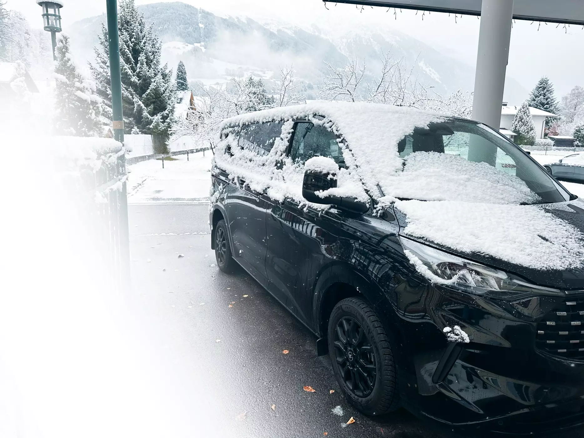 Verschneiter Ford Bus in schwarzer Lackierung vor einer Hoteleinfahrt. Im Hintergrund verschneite Berge.